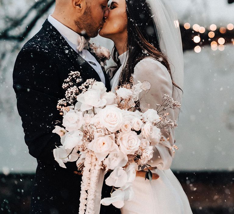 Bride and groom share a kiss at their snowy wedding with the bride holding a large white winter wedding bouquet 