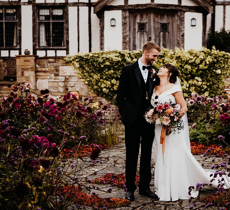 Bride in off the shoulder wedding dress gazing up at groom in black tuxedo in front of the Hertfordshire wedding venue, The Barn at Alswick 