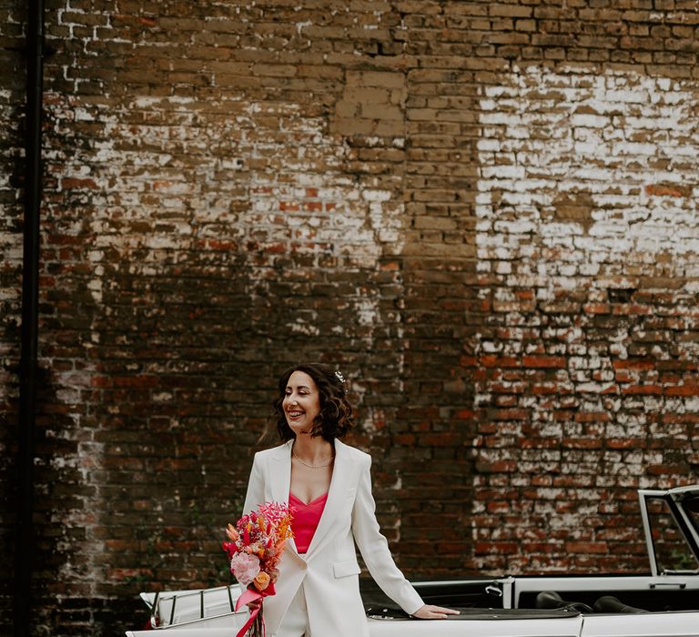 The stylish modern bride gets ready to leave for her retro styled wedding ceremony with a white retro wedding car and hot pink bouquet 