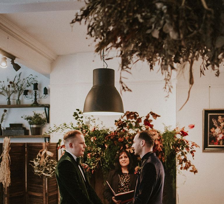 Wedding flowers at autumnal spooky wedding with the two grooms reading out their wedding vows wearing luxe velvet suit jackets