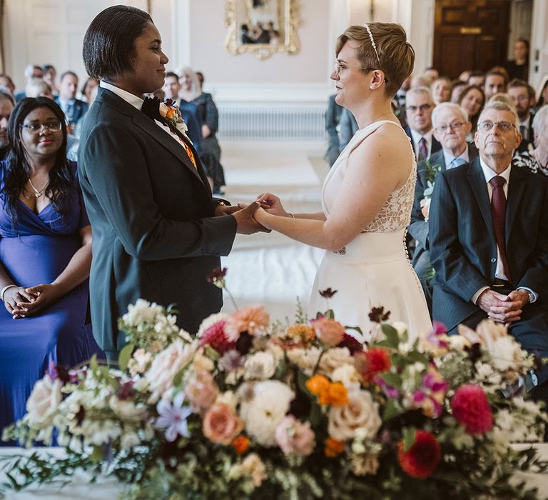 Bride in black suit jacket with the bride in a white wedding dress holding hands for their same sex lesbian wedding ceremony