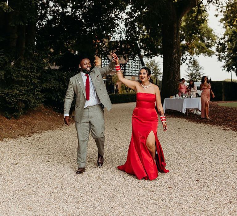 Groom in all grey wedding suit holds hands with the bride in a long strapless bright red after party outfit change dress for Larmer Tree Gardens wedding 