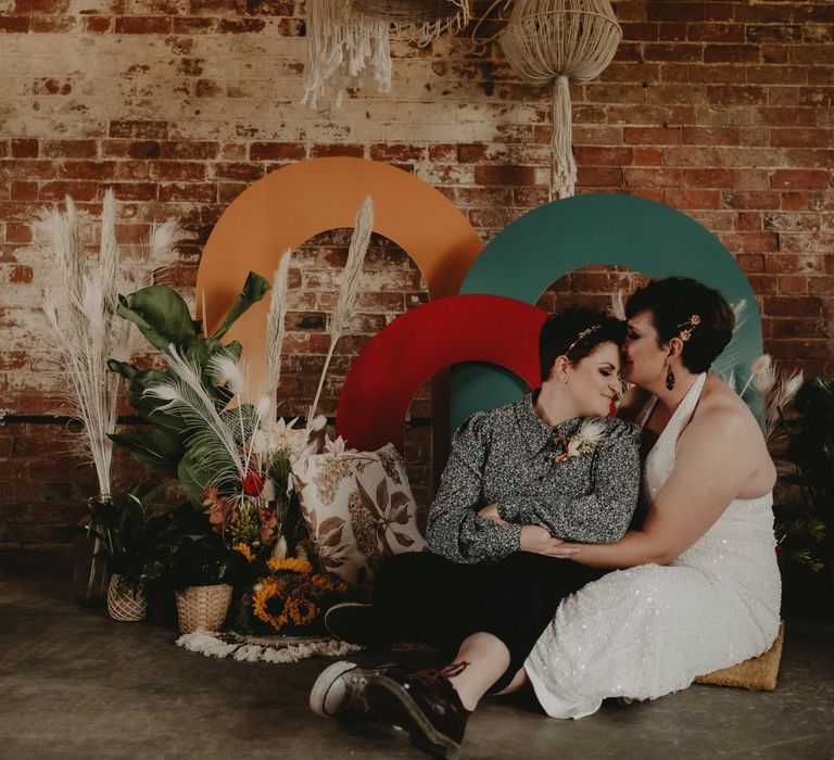 Two brides sitting in front of a colourful backdrop 