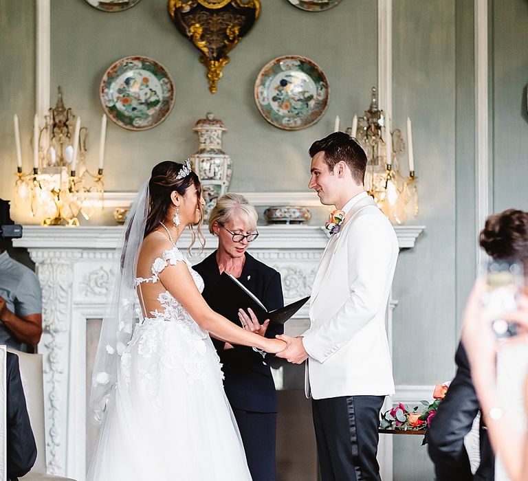 Bride in an off the shoulder Galia Lahav wedding dress with lace detail holding hands with her groom in a white tuxedo jacket at Leeds castle 