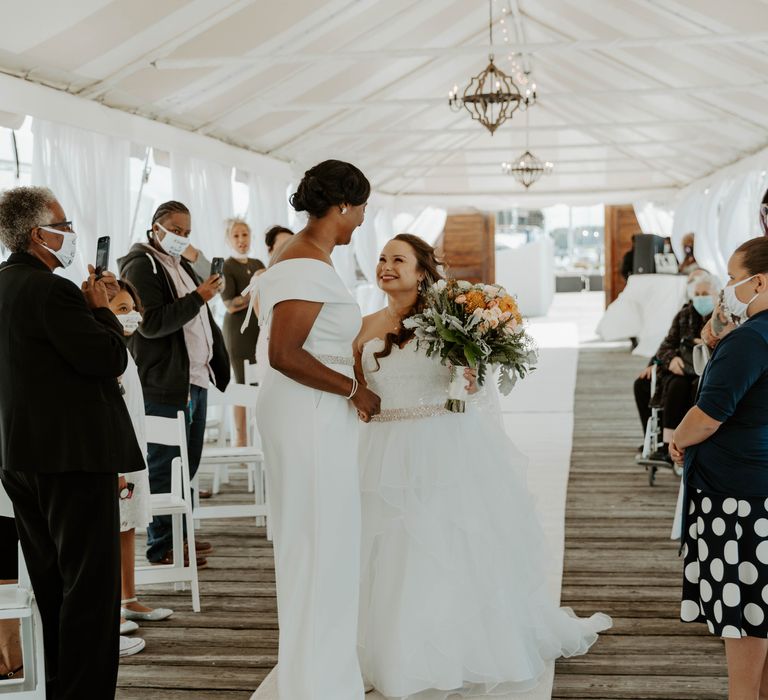 Black bride in an off the shoulder jumpsuit meets her bride in a strapless wedding dress as she walks down the aisle unaccompanied