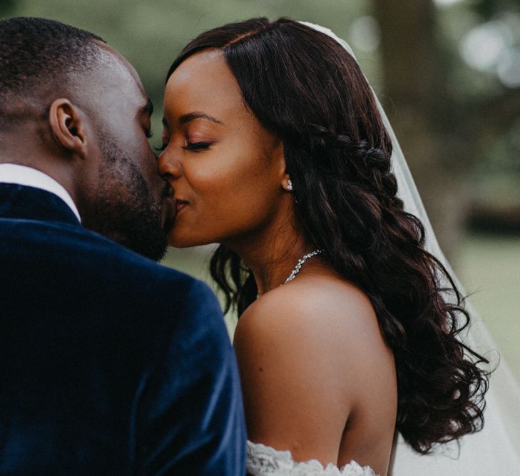 Wedding hairstyles for Black brides. A Black bride and groom kiss. The bride has long loose curls and wears a veil.