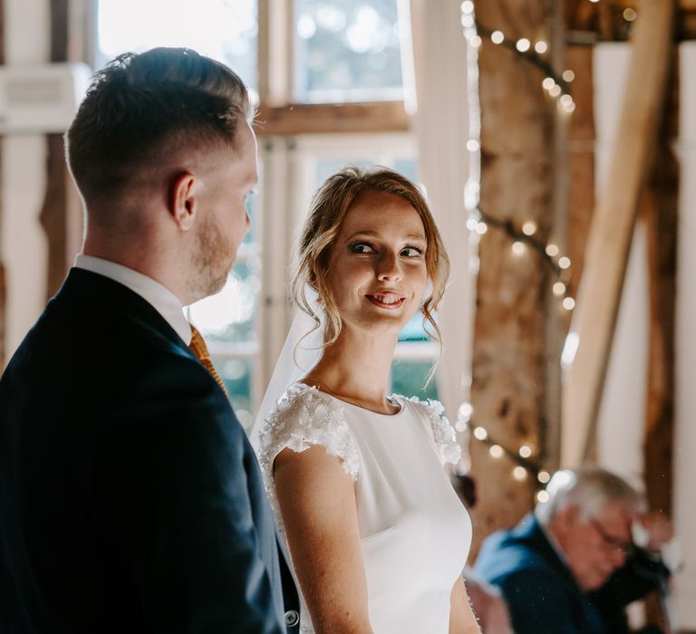 Red headed bride in a fitted Pronovias wedding dress with appliqué shoulder detail standing at the altar with her groom 