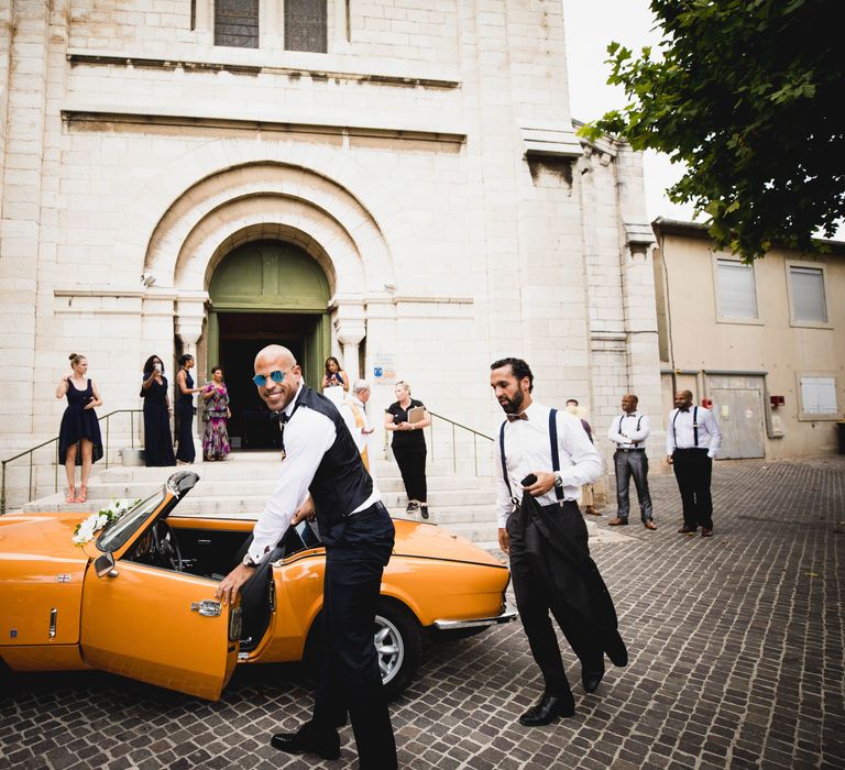 The groom arriving at the church in a vintage orange open top sports car