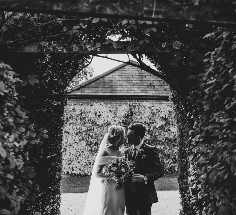 Black & white image of bride and groom kissing under green foliage arch