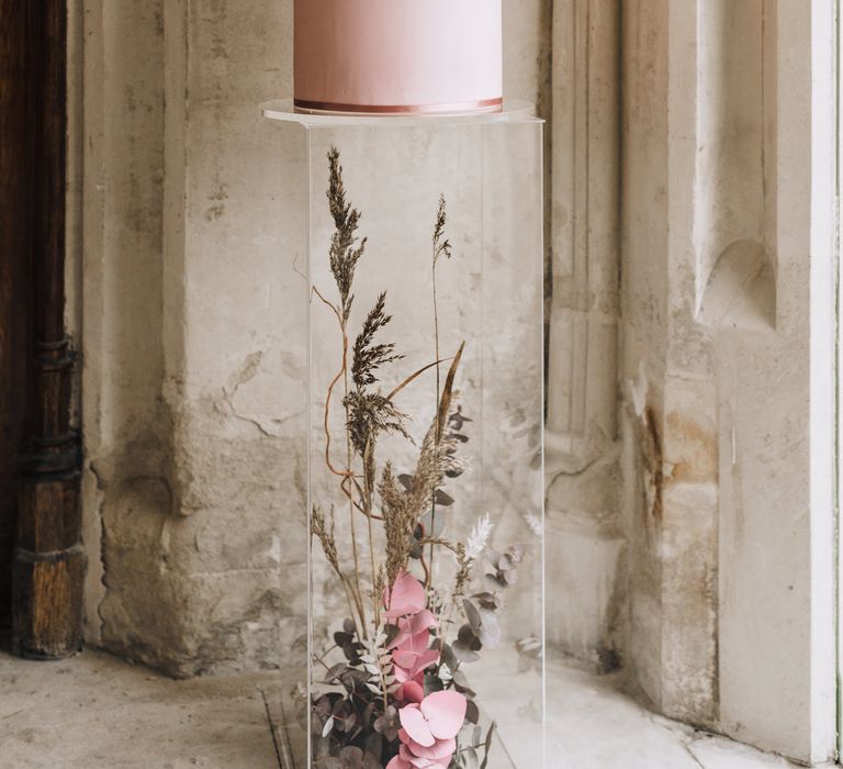 Blush pink four tier wedding cake with flower design standing on an acrylic plinth filled with decorative flowers. 