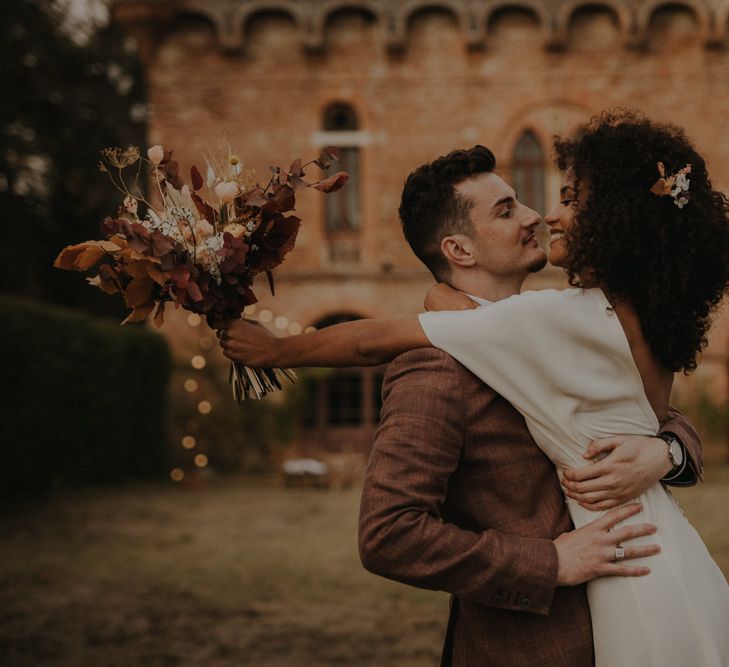 Groom in a brown check suit picking up his bride in a crepe wedding dress with naturally curly hair holding an autumn leaf wedding bouquet 