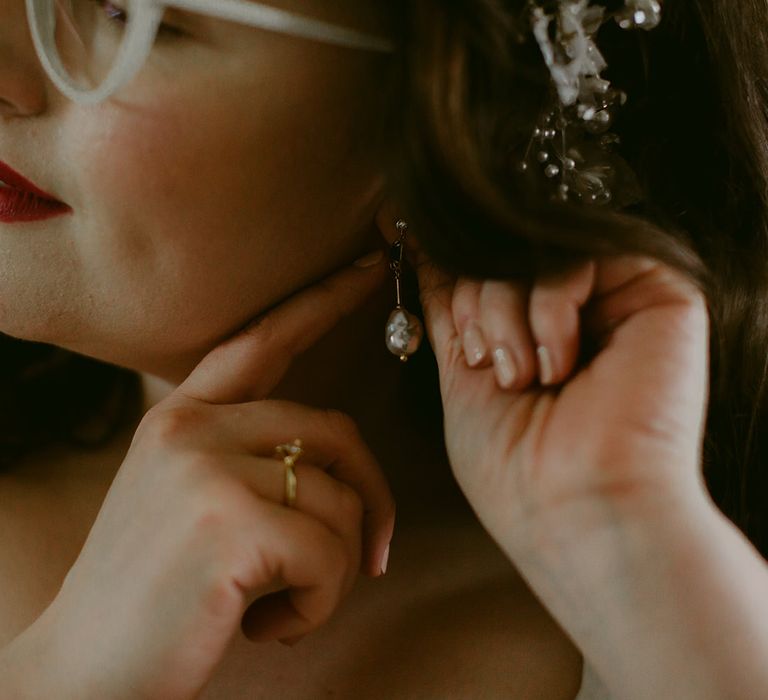 Bride with bold red lip and white framed glasses puts in gold drop earrings