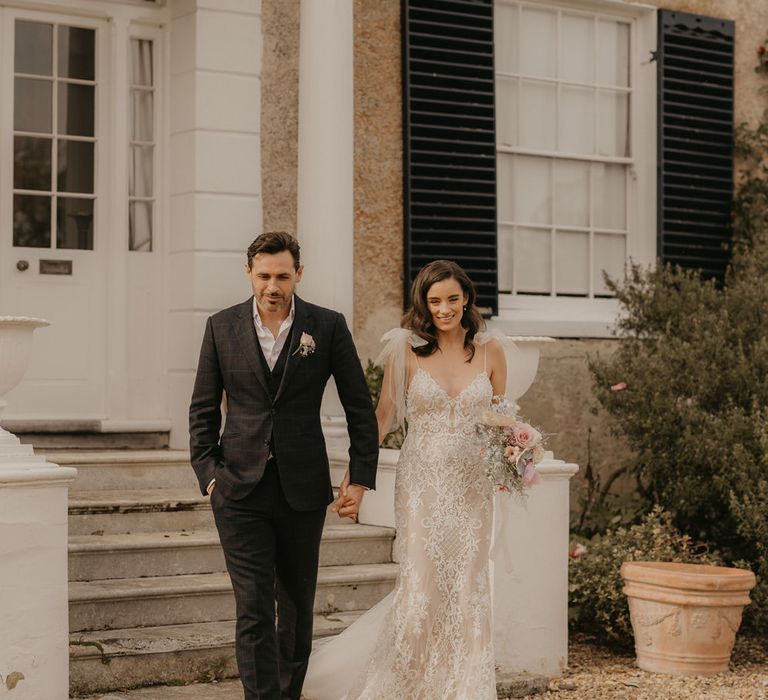 Groom in a dark check suit and tan monk shoes holding hands with his bride in a fitted lace wedding dress with thin straps 