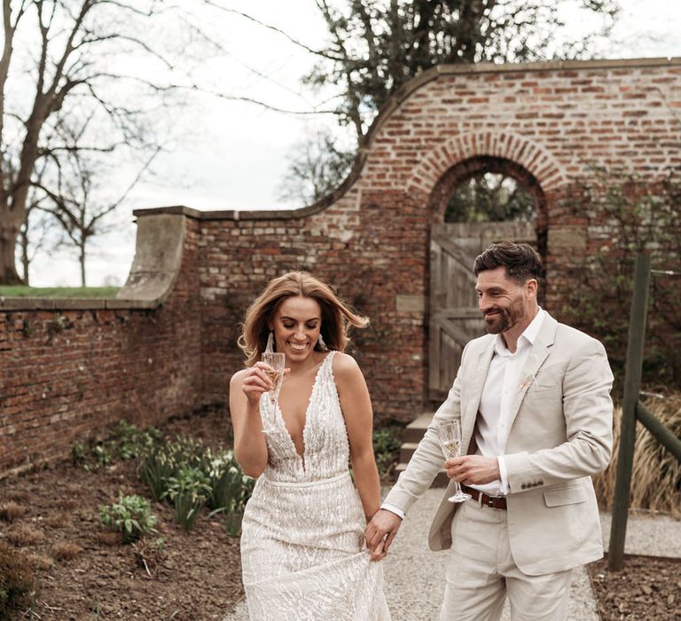 Groom in a sand suit holding hands with his bride in a plunging neck wedding dress drinking champagne 