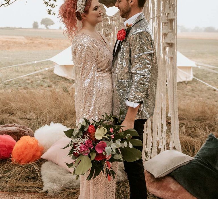 Bride in long sleeved gold sequinned wedding dress and beaded crown headband stands facing groom in silver sequin suit jacket and red flower buttonhole in front of macrame hanging outdoors at festival themed wedding