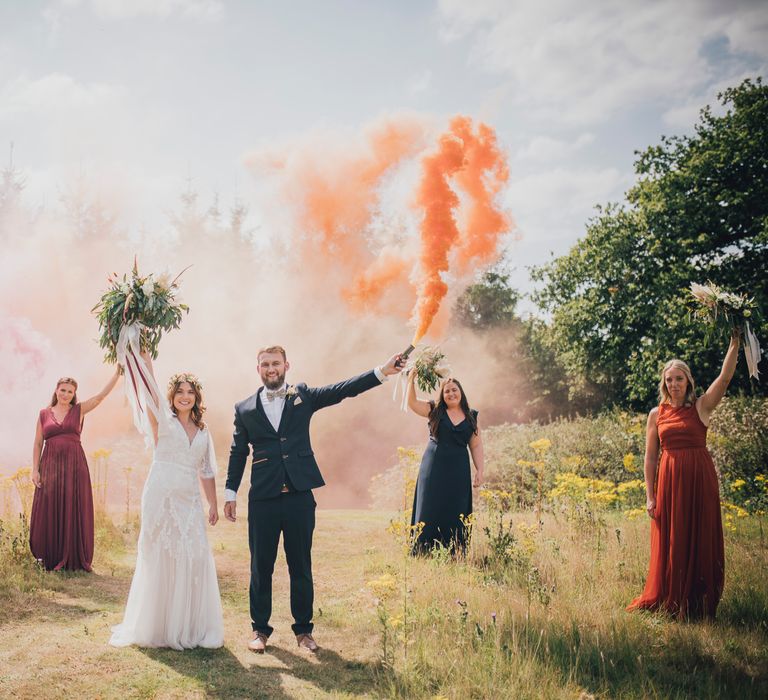 Bride & groom stand in field surrounded by wedding party and smoke bombs