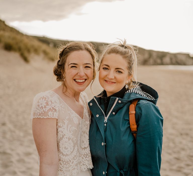 Smiling bride in white lace top wedding dress with capped sleeves and satin skirt stands with photographer in blue ran jacket on the beach after Dunluce Castle wedding