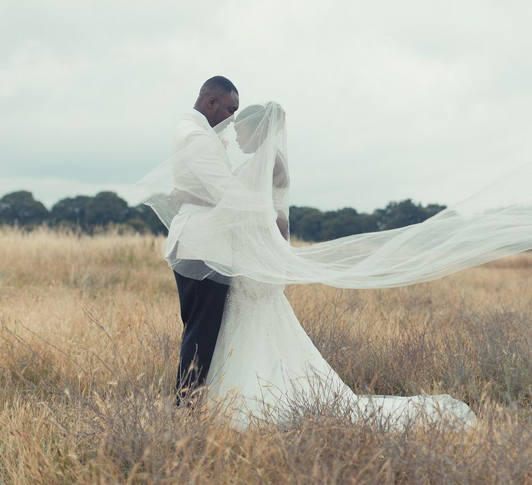 Bride & groom stand outdoors and brides veil blows around them in the countryside 