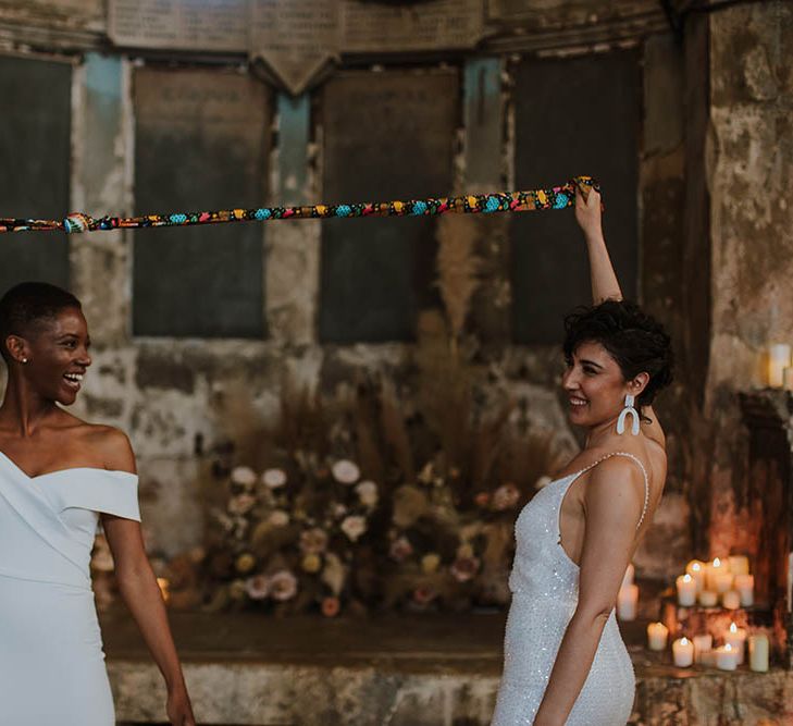 Two brides holding their hand fasting knot in the air at The Asylum wedding venue with pillar candles decorating the altar 