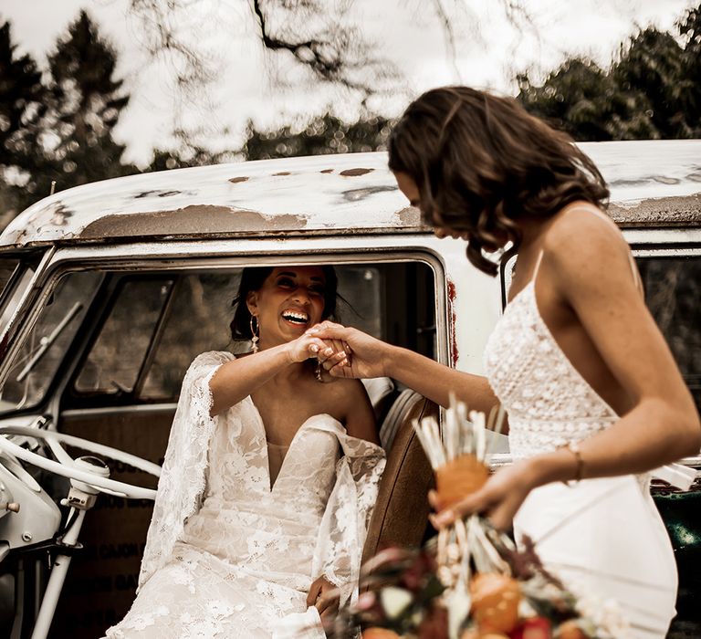 Bride in a fitted wedding dress with thin straps helping her bride out of the car in a boho wedding dress with lace batwings 