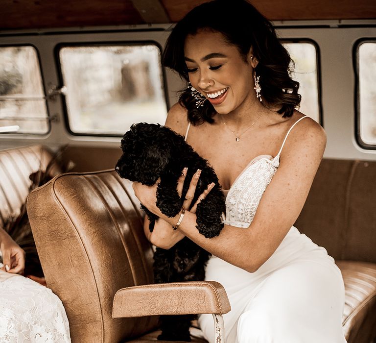 Black bride in a fitted wedding dress with thin straps holding a black cockapoo puppy in a VW camper van 