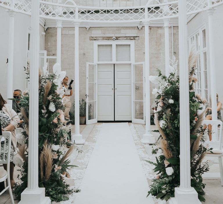 Interior of glasshouse at Came House Dorset with white pillars, white carpeted aisle and green and white floral installations