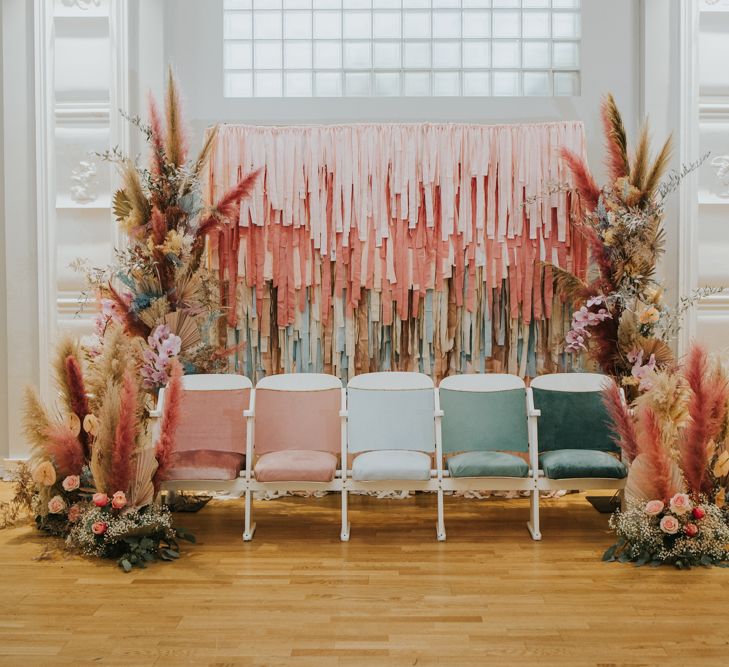 A row of velvet pastel coloured wedding chairs in front of a tissue paper backdrop flanked by vertical flower arrangements with pampas grass and dried palm leaves 