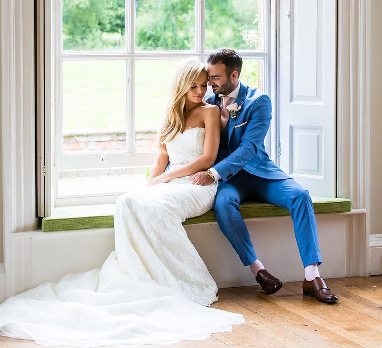 Blonde white bride in white dress sits at window seat with white groom in bright blue suit