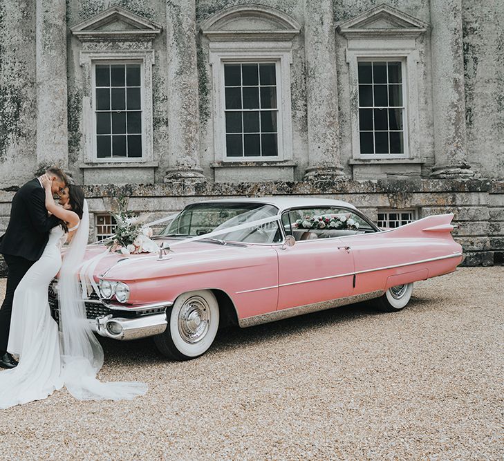 Bride in white Pronovias wedding dress with train and veil holds hands on grooms chest as they lean on pink Cadillac in front of Came House Dorset after their wedding