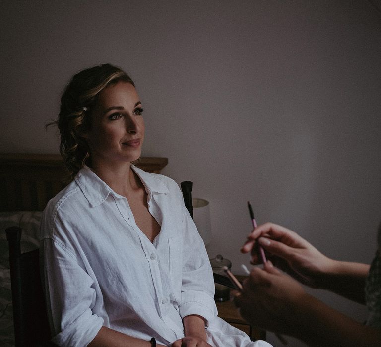 Bride with curled wedding updo in oversized white shirt sits whilst having her make up done before garden wedding with burnt orange wedding theme