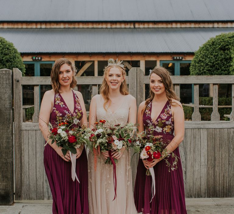 Bride in pearl wedding dress and bridesmaids in burgundy embroidered bridesmaids dresses stand outside all holding red and green wedding bouquets after barn wedding ceremony 