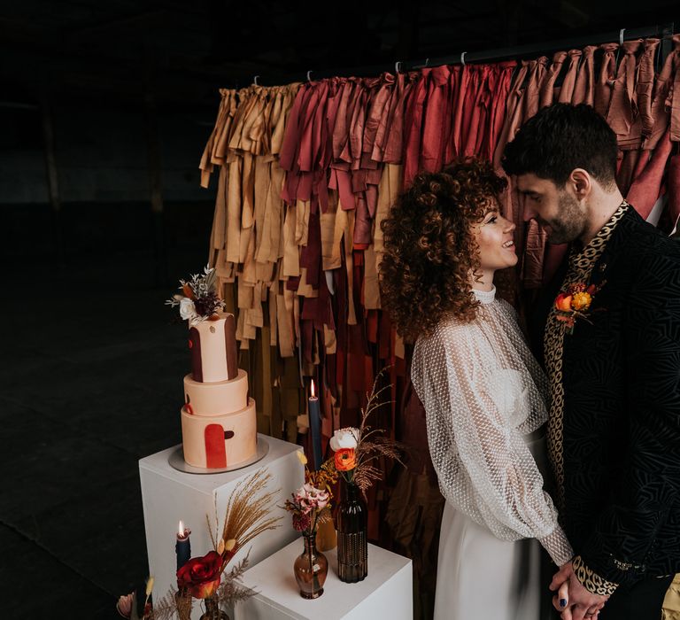 Bride and groom standing in front of an ombre fabric backdrop with retro cake, bottles and flower decor 