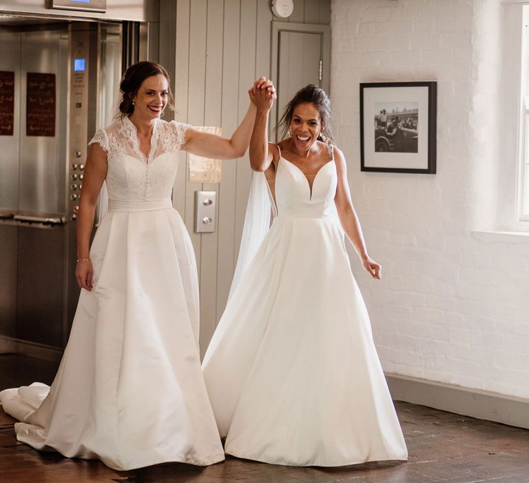 Bride in white cami wedding holds up hand of bride in white lace top capped sleeve wedding dress as they exit lift into wedding reception at The West Mill Derby