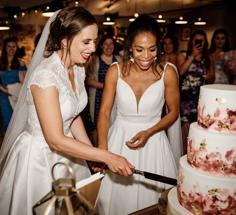 Smiling bride in white lace top capped sleeve weeding Dres and veil cuts three tiered wedding cake whilst holding hands with bride in white cami wedding dress with should veil details during wedding reception at The West Mill Derby