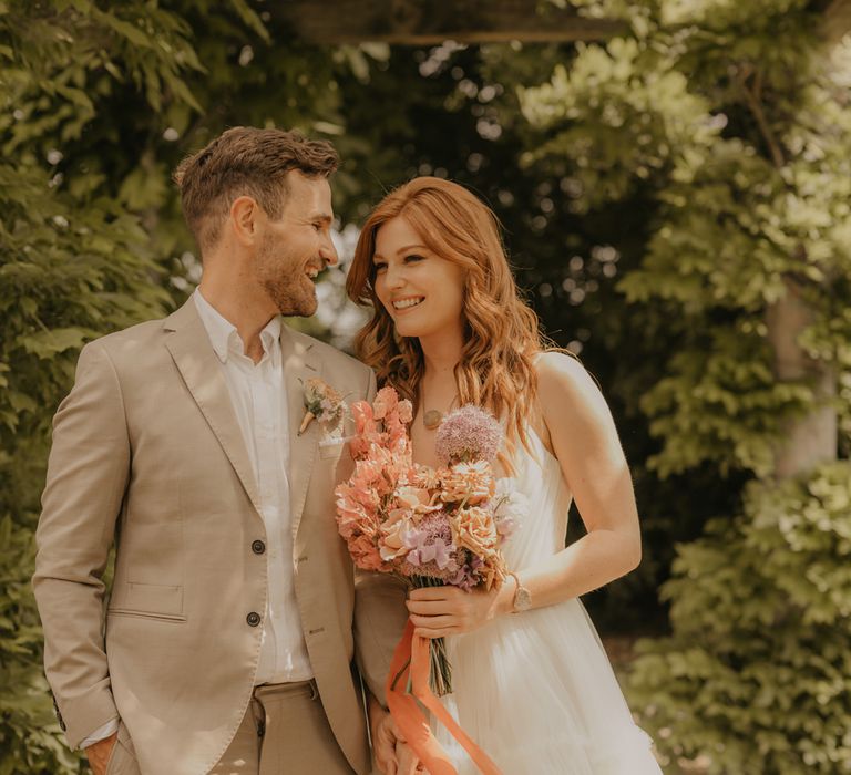 Groom in a tan suit laughing with his bride in a Made With Love wedding dress as she holds a pastel orange and pink bouquet tied with ribbon 