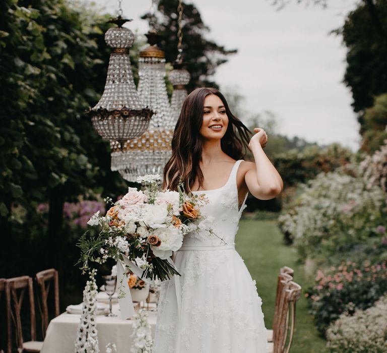 Beautiful bride in an appliqué wedding dress with full skirt, fitted bodice and straps standing an an outdoor tablescape with chandelier installation