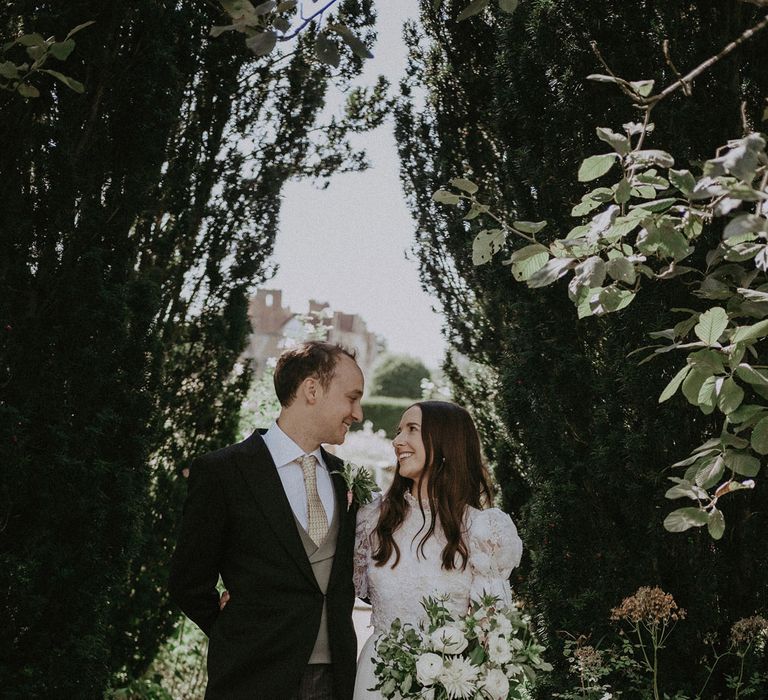 Bride in white lace puffed sleeve Daalarna wedding dress holding white and greenn bridal bouquet stands arm in arm with groom in morning coat and green waistcoat in grounds of Loseley Park after summer wedding in Surrey