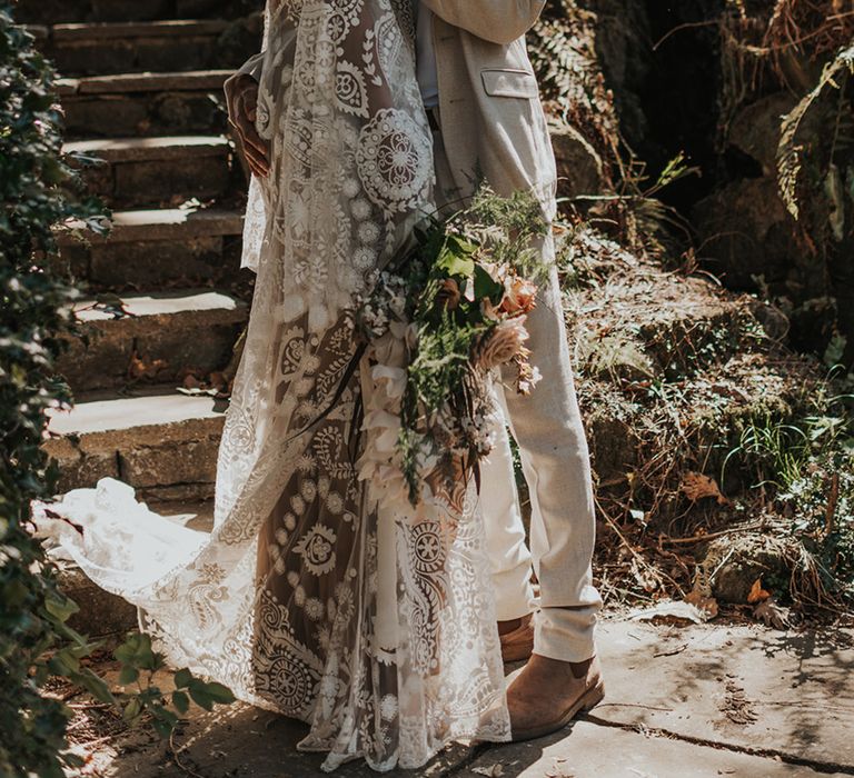 Groom in a beige suit and grandad collar shirt embracing his bride in a lace wedding dress