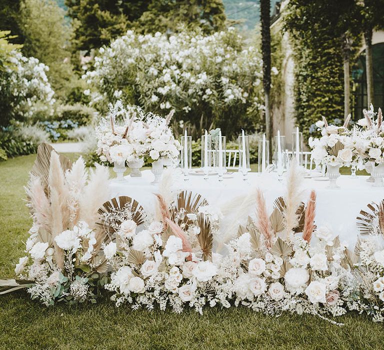 Beautifully elegant styled tablescape complete with floral installations in white and neutral tones with green foliage and pampas grass outdoors