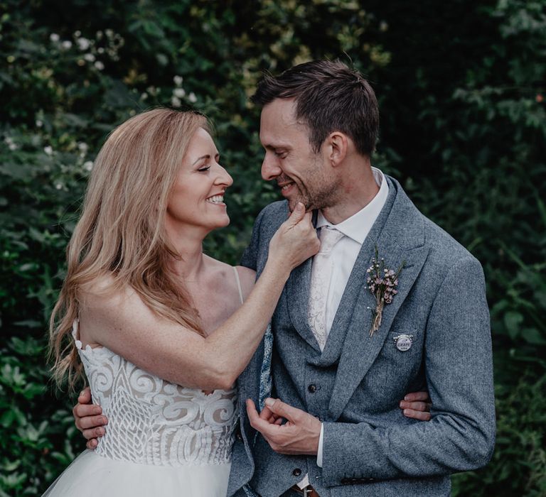 Bride in a tulle skirt wedding dress touch her grooms chin in a grey wool wedding suit 