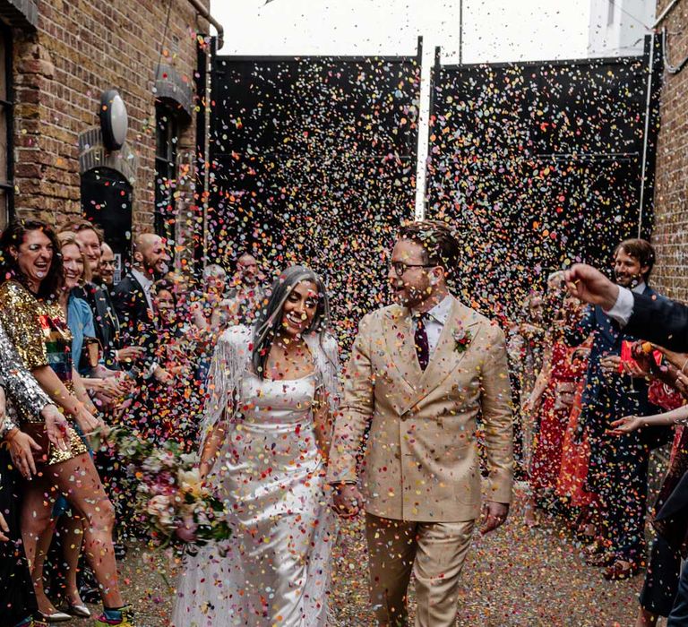 Bride in satin Halfpenny London wedding dress and tasselled bridal cape and groom in double breasted linen suit and gold Nike trainers walk through multicoloured confetti thrown by guests for wedding at Loft Studios London