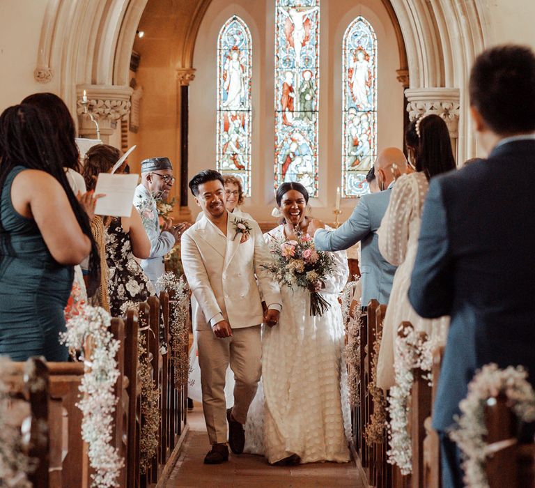 Bride in long mesh sleeve spotted Vagabond wedding dress holding mixed bridal bouquet and groom in double breasted linen suit walk down the aisle after church wedding ceremony at Wasing Park