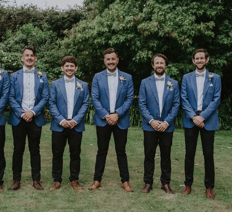 Groom and groomsmen stand in garden wearing blue suit jackets, white shirts, sage green bow ties and floral buttonholes at Isle of Wight wedding with macrame wedding decor