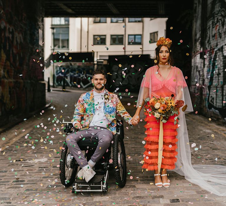 Tetraplegic disabled groom in grey trousers and colourful blazer holding hands with his bride in a coral wedding dress and flower crown 