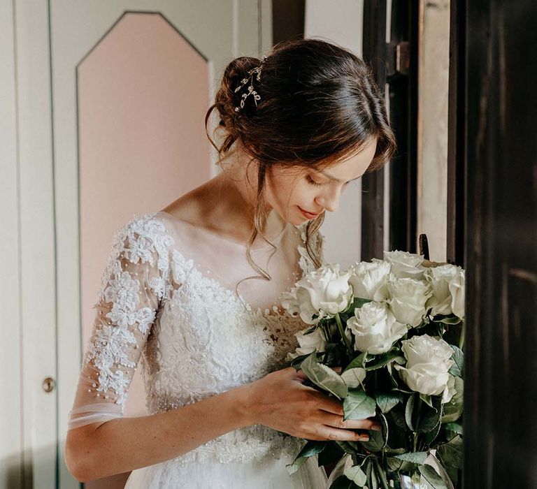 Bride wears her dark hair pulled back into low bun as curled tendrils curl around her face and she looks down at white floral bouquet with green foliage  | Royal Studio