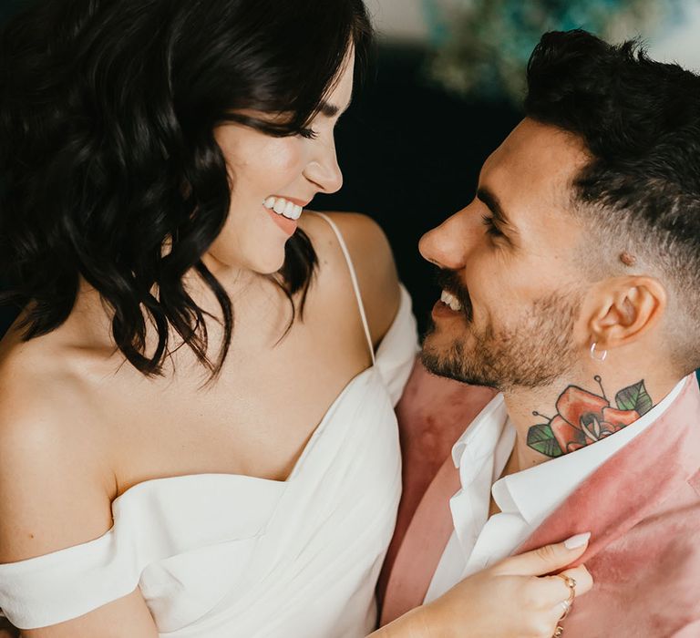 Beautiful bride with shoulder length wavy hair in a cold shoulder wedding dress sitting on her grooms lap in a pink velvet jacket 