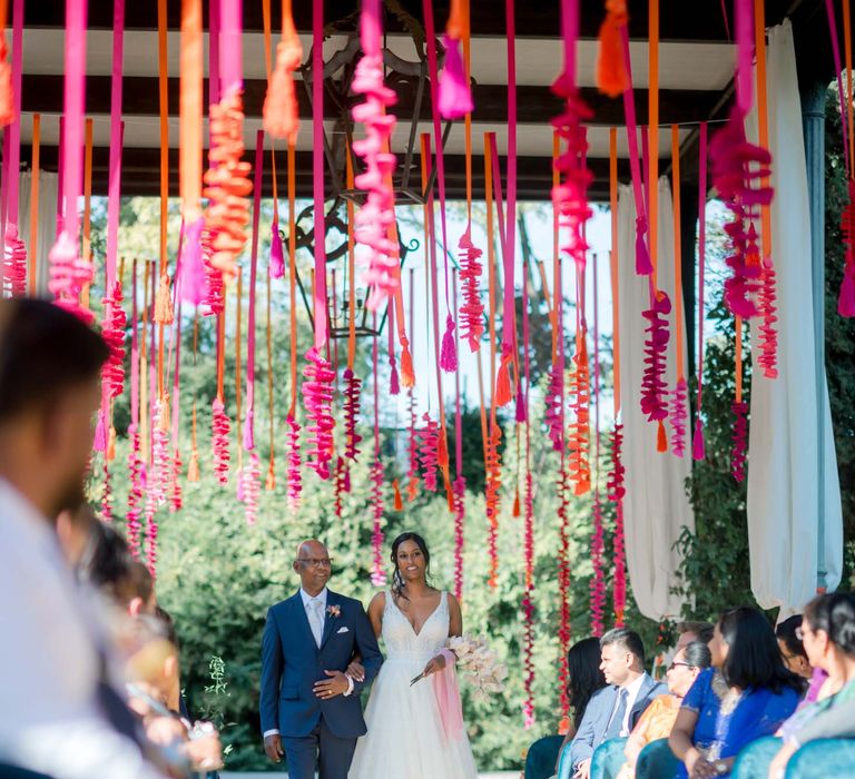 Bride walks down the aisle with her father whilst colourful hot pink and orange hangings suspend from the ceiling 