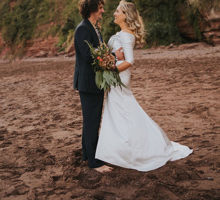 Bride & groom look lovingly at one another as they stand on the beach