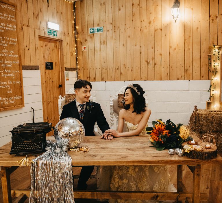 Bride in strapless wedding dress and headpiece sits with groom in dark blu suit and floral tie during rustic wedding ceremony