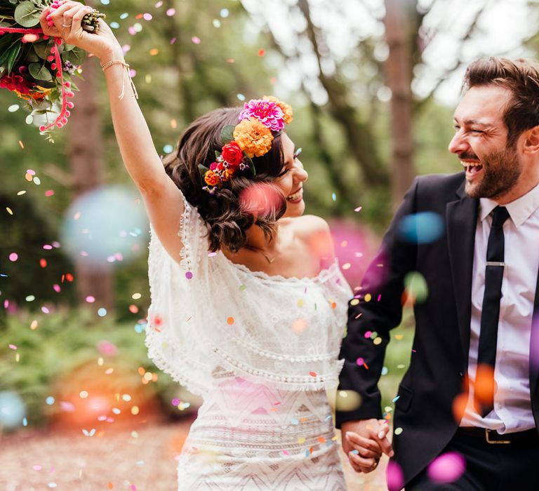 Bride in off the shoulder Grace Loves Lace wedding dress ad colourful flower crown holding multicoloured bridal bouquet holds hands with groom in black suit under multicoloured confetti after outdoor wedding ceremony